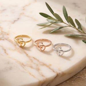 May include: Three rings in gold, rose gold, and silver tones, each featuring a stylized letter design. The rings are arranged in a row, showcasing a polished metallic finish and a thin, circular band structure for each individual piece.
