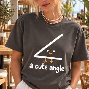 Puede incluir: Una camiseta de manga corta gris carbón con un gráfico blanco de un ángulo agudo con cara sonriente y pies naranjas. Debajo del gráfico, el texto a cute angle aparece en letras minúsculas blancas. La camiseta tiene cuello redondo.
