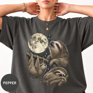 以下が含まれることがあります： 大きな満月と3匹のナマケモノが描かれたチャコールグレーのTシャツ。1匹は枝にぶら下がり、もう1匹は上を見上げ、3匹目は枝の上で休んでいます。左下には白い文字でPEPPERとプリントされています。