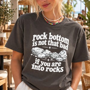 以下が含まれることがあります： チャコールグレーの半袖Tシャツ。中央には「rock bottom is not that bad if you are into rocks」という白い文字と、いくつかの岩を描いたグラフィックがプリントされています。クルーネックでゆったりとしたフィット感のTシャツです。