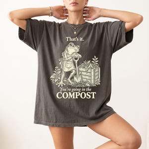 Può includere: Una t-shirt grigio antracite con la grafica di una rana che indossa un grembiule, appoggiata a una vanga accanto a una compostiera in legno. Il testo sopra recita "That's it." e il testo sotto "You're going in the COMPOST" in bianco.