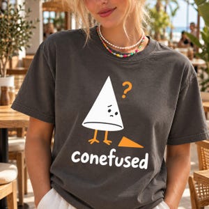 May include: A charcoal grey short-sleeve t-shirt featuring a graphic of a white cone character with orange legs and a sad expression. An orange question mark is above the cone, and the word "conefused" is printed in white below the graphic.