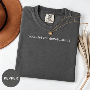 Op de afbeelding: Een antracietgrijs T-shirt met korte mouwen en de witte tekst "bark-driven development" op de borst. Het shirt is voorzien van een Comfort Colors-merklabel bij de binnenhalslijn en heeft maat S.