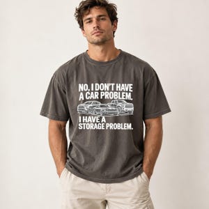 Puede incluir: Una camiseta de manga corta gris carbón con texto en blanco que dice: "NO, I DON'T HAVE A CAR PROBLEM. I HAVE A STORAGE PROBLEM." El diseño incluye ilustraciones de líneas blancas de tres vehículos diferentes situadas entre las líneas de texto.
