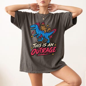 Könnte beinhalten: Ein anthrazitfarbenes Kurzarm-T-Shirt mit einer Grafik eines blauen Dinosauriers, auf dessen Rücken eine braune Katze mit Sonnenbrille und Krone reitet. Der Text "THIS IS AN OUTRAGE" ist darunter in fetten rosa und weißen Buchstaben mit Sternen gedruckt.
