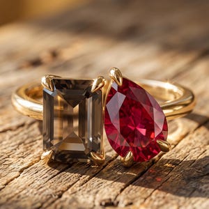 Op de afbeelding: Een gouden ring met twee opvallende edelstenen naast elkaar. De ene steen is donkergrijs en smaragdgeslepen, de andere is felrood en peervormig. Beide stenen zijn gezet in gouden klauwen op een gepolijste gouden ringband.