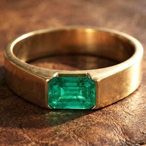 Op de afbeelding: Een gouden bandring met een enkele smaragdgeslepen groene edelsteen die verzonken in het midden van het metaal is geplaatst. De ring heeft een gladde, gepolijste afwerking en een brede, massieve band die de rechthoekige steen stevig vasthoudt.