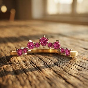 Könnte beinhalten: Ein goldener Ring mit einer geschwungenen Ringschiene, die mit acht runden, leuchtend pinkfarbenen Edelsteinen besetzt ist. Die Steine sind in einem abgestuften Bogen angeordnet, wobei der größte Stein in der Mitte sitzt und von kleinen Metallkrappen gehalten wird.