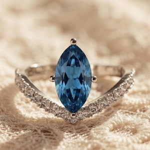 Op de afbeelding: Een zilverkleurige ring met een grote, blauwe edelsteen in marquise-slijpvorm in het midden. De ringband is V-vormig en versierd met een rij kleine, heldere, rond geslepen steentjes over de gehele lengte, wat zorgt voor een sprankelend ontwerp.