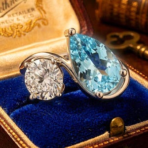 Könnte beinhalten: Ein Bypass-Ring mit einem runden, im Brillantschliff geschliffenen, klaren Stein und einem hellblauen, birnenförmigen Stein, gefasst in eine polierte, silberfarbene Metallschiene. Die beiden Steine liegen nebeneinander und bilden ein asymmetrisches Design.