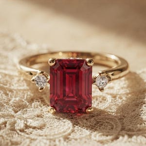 Op de afbeelding: Een gouden ring met een grote, rechthoekige, smaragdgeslepen dieprode edelsteen in het midden, geflankeerd door twee kleine, ronde, helderwitte stenen aan weerszijden. De ringband is van gepolijst geelgoud met een klassiek ontwerp met drie stenen.