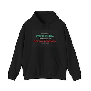 Op de afbeelding: Een zwarte hoodie met een grafische print met de tekst: ENVIRONMENT: Works in dev. Production? Not my problem. De tekst wordt weergegeven in een combinatie van groene, witte en rode kleuren tegen een donkere rechthoekige achtergrond.