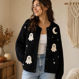 May include: A black long-sleeve button-up cardigan featuring white crocheted appliques of ghosts, stars, and a crescent moon. The garment has a round neckline, ribbed cuffs, and a textured knit pattern, worn over a white tank top and blue jeans.