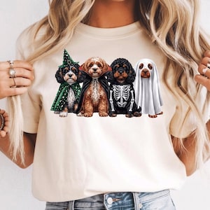 May include: A cream-coloured t-shirt featuring a graphic print of four dogs in Halloween costumes. The dogs are dressed as a wizard in a green star-patterned cape, a vampire in a black cape, a skeleton, and a ghost under a white sheet.