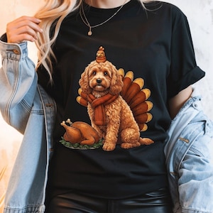 Könnte beinhalten: Ein schwarzes T-Shirt mit einer grafischen Illustration eines goldfarbenen, lockigen Hundes, der einen Schal und einen kleinen Vogel auf dem Kopf trägt. Hinter dem Hund ist ein Truthahn-Schwanzfächer und ein gebratener Truthahn auf grünem Grund.