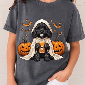 Könnte beinhalten: Ein anthrazitfarbenes Kurzarm-T-Shirt mit einer Grafik eines schwarzen, lockigen Hundes in einem cremefarbenen Geisterkostüm. Der Hund hält ein orangefarbenes Getränk mit Geister-Symbol, flankiert von zwei orangefarbenen Kürbissen und kleinen Fledermäusen.