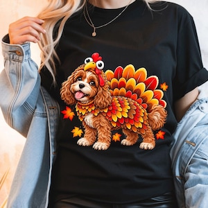 May include: A black short-sleeved t-shirt featuring a graphic of a brown spaniel dog wearing a turkey costume with a red wattle, a beak, and a colourful fan of orange, yellow, and red feathers, surrounded by small orange autumn leaves.