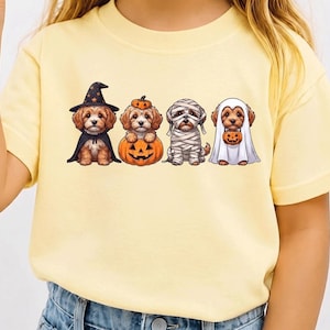 Könnte beinhalten: Ein hellgelbes Kurzarm-T-Shirt mit einem Grafikdruck von vier braunen Welpen in Halloween-Kostümen. Die Welpen sind als Hexe, Kürbis, Mumie und Geist verkleidet, wobei ein Welpe einen kleinen Kürbiseimer als Süßigkeitenbehälter hält.