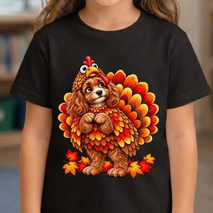May include: A black short-sleeved t-shirt featuring a graphic of a brown spaniel dog wearing a turkey costume with a red and yellow feathered tail and a turkey head hood, surrounded by scattered orange and red autumn maple leaves.