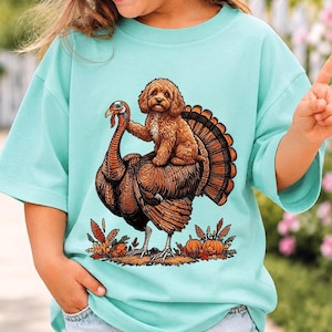 以下が含まれることがあります： 明るいミントグリーンの半袖Tシャツ。大きな七面鳥の背中に乗った茶色の巻き毛の犬のグラフィックプリントが施されています。デザインの足元には、オレンジ色のカボチャや落ち葉など、秋をテーマにした要素が描かれています。