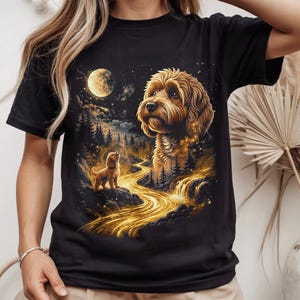Op de afbeelding: Een zwart T-shirt met korte mouwen en een grafische print van een goudkleurige hond die opkijkt naar een volle maan in een sterrenhemel. De illustratie toont een gloeiende gouden rivier die door een donker boslandschap stroomt met een kleine hond.