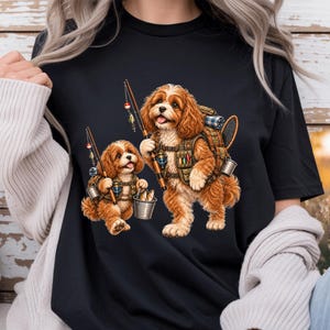 Könnte beinhalten: Ein schwarzes T-Shirt mit einer grafischen Illustration von zwei braunen, flauschigen Hunden in Angelausrüstung. Die Hunde halten Angelruten, tragen Westen mit Taschen und haben einen Metalleimer bei sich, der mit kleinen Fischen gefüllt ist.
