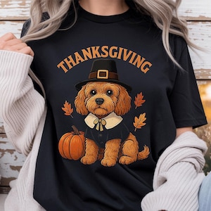 May include: A black t-shirt featuring a graphic of a brown curly-haired dog wearing a black pilgrim hat and a white collar. The dog sits next to an orange pumpkin, surrounded by orange autumn leaves. The word THANKSGIVING is printed in orange above.