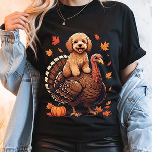 Puede incluir: Una camiseta negra de manga corta con un estampado gráfico de un cachorro marrón claro sentado sobre el lomo de un pavo marrón. El diseño incluye hojas de arce otoñales naranjas y rojas dispersas y una calabaza naranja en la parte inferior.