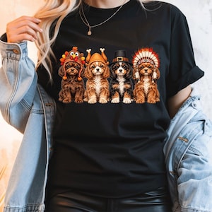 May include: A black short-sleeved t-shirt featuring a graphic print of four brown and white puppies sitting in a row. Each puppy wears a different headpiece, including a turkey hat, a cooked turkey hat, a black pilgrim hat, and a feathered headdress.