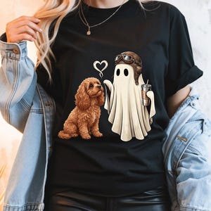 May include: A black t-shirt featuring a graphic print of a brown curly-haired dog sitting next to a white ghost wearing a brown aviator cap and goggles. The ghost is holding a coffee cup, and a small white heart outline is positioned above them.