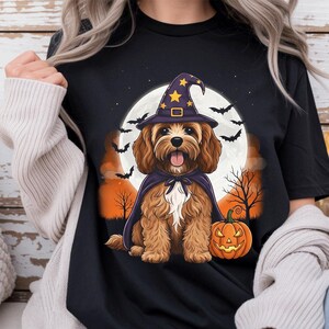 May include: A black t-shirt featuring a graphic of a brown dog wearing a purple witch hat with yellow stars and a matching cape. The dog sits in front of a large white full moon, orange clouds, black silhouettes of bats, trees, and a jack-o'-lantern.
