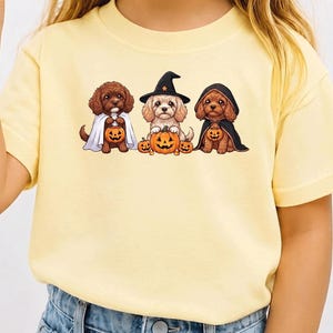 Puede incluir: Una camiseta de manga corta de color amarillo pálido con un estampado gráfico de tres perros pequeños con disfraces de Halloween. Los perros llevan una capa de fantasma blanca, un sombrero de bruja negro y una capa con capucha negra y calabazas.