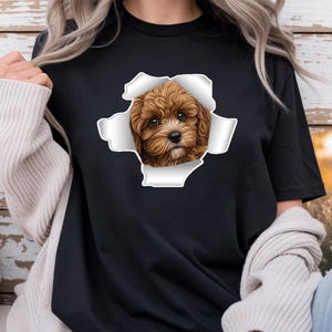 Puede incluir: Una camiseta negra de manga corta con un estampado gráfico de un cachorro marrón y esponjoso que asoma a través de un diseño de papel blanco rasgado. El cachorro tiene ojos grandes y oscuros y un pelaje detallado y texturizado en el centro.