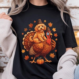 Peut inclure: Un t-shirt noir à manches courtes avec une impression graphique représentant un chiot doré assis sur le dos d'une dinde. Le chiot et la dinde portent tous deux de petits chapeaux de pèlerin marron. Des feuilles d'automne orange et une citrouille.