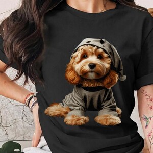 May include: A black short-sleeved t-shirt featuring a graphic print of a fluffy golden-brown puppy wearing a grey hooded pajama set. The puppy has large dark eyes and is positioned in the center of the shirt, facing forward with a calm expression.