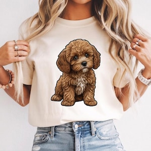 Peut inclure: Un t-shirt couleur crème arborant une illustration graphique centrée d'un chiot brun au pelage bouclé, aux grands yeux sombres et avec une petite tache blanche sur le poitrail. Le chiot est représenté assis avec un contour noir.