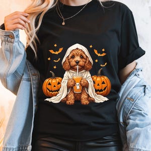 May include: A black t-shirt featuring a graphic of a brown curly-haired dog wearing a white ghost costume. The dog holds an orange drink with a ghost icon. Two orange jack-o'-lanterns and several small orange bats surround the dog on the shirt.