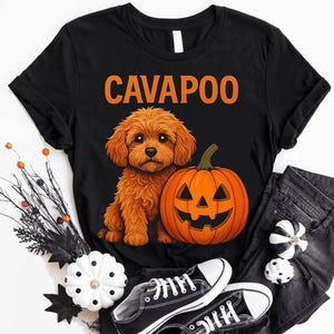 May include: A black short-sleeved t-shirt featuring a graphic of a brown Cavapoo dog sitting next to an orange jack-o'-lantern pumpkin. The word CAVAPOO is printed in large orange capital letters above the dog and pumpkin illustration.