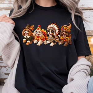 May include: A black short-sleeved t-shirt featuring a graphic print of four small dogs in a row. The dogs are dressed in autumn-themed costumes, including a pilgrim hat, a turkey outfit, a feathered headdress, and a cornucopia with sunflowers.