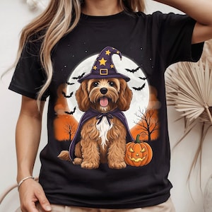 Peut inclure: Un t-shirt noir avec une illustration d'un chien marron portant un chapeau de sorcière violet à étoiles jaunes et une cape assortie. Le chien est assis à côté d'une citrouille orange sculptée devant une pleine lune, des nuages orange, des arbres nus et des chauves-souris noires.
