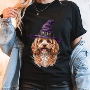 May include: A black short-sleeved t-shirt featuring a graphic print of a curly-haired brown dog wearing a purple witch hat with a dark belt and a crescent moon buckle. The dog has a light-colored chest, floppy ears, and an open mouth with a pink tongue.