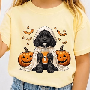 Op de afbeelding: Een lichtgeel t-shirt met een afbeelding van een zwarte puppy in een wit spookkostuum, die een oranje drankje met een spookicoontje vasthoudt. De puppy wordt geflankeerd door twee oranje pompoenen en kleine oranje vleermuizen erboven.