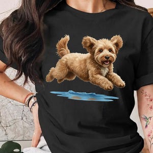 May include: A black short-sleeved t-shirt featuring a graphic print of a light brown, fluffy dog jumping over a small blue puddle of water. The dog has perky ears, a happy expression with its mouth open, and a white patch of fur on its chest.