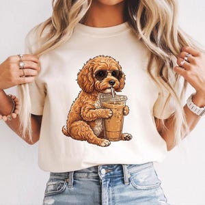 May include: A cream-coloured short-sleeve t-shirt featuring a graphic illustration of a curly brown dog wearing black sunglasses and holding a clear plastic cup filled with an iced coffee beverage with a straw, centred on the front of the shirt.