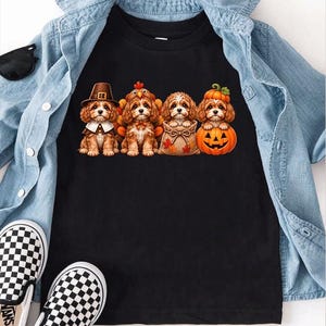 May include: A black t-shirt featuring a graphic of four brown puppies. One puppy wears a pilgrim hat, one wears a turkey costume, one sits in a burlap sack, and one sits inside a carved orange pumpkin. The shirt is layered under a light blue denim shirt.