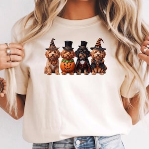 Könnte beinhalten: Ein cremefarbenes T-Shirt mit einem Grafikdruck von vier Hunden in Halloween-Kostümen. Die Hunde tragen verschiedene Hüte, einen Kürbisanzug, einen Vampirumhang und ein gestreiftes Kleid, die in einer Reihe auf der Brust angeordnet sind.