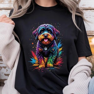 Può includere: Una t-shirt nera con una vivace grafica multicolore raffigurante un cucciolo. L'illustrazione del cane utilizza una tavolozza di colori arcobaleno con effetti di schizzi di vernice attorno alle zampe. Il cucciolo indossa un collare scuro.