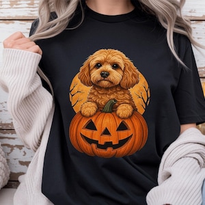 Puede incluir: Una camiseta negra con una ilustración gráfica de un cachorro de color marrón dorado y pelaje esponjoso sentado dentro de una calabaza naranja tallada. El fondo del gráfico muestra una gran luna llena amarilla con siluetas de ramas desnudas.