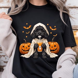 May include: A black t-shirt featuring a graphic of a black puppy wearing a beige hooded cloak, holding an orange drink with a ghost icon. The puppy is flanked by two orange jack-o'-lanterns, with several small orange bats flying above.