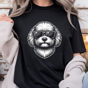 May include: A black short-sleeved t-shirt featuring a central graphic illustration of a fluffy dog wearing dark sunglasses. The dog is depicted in a detailed black and white line art style with a white outline, set against the solid black fabric.